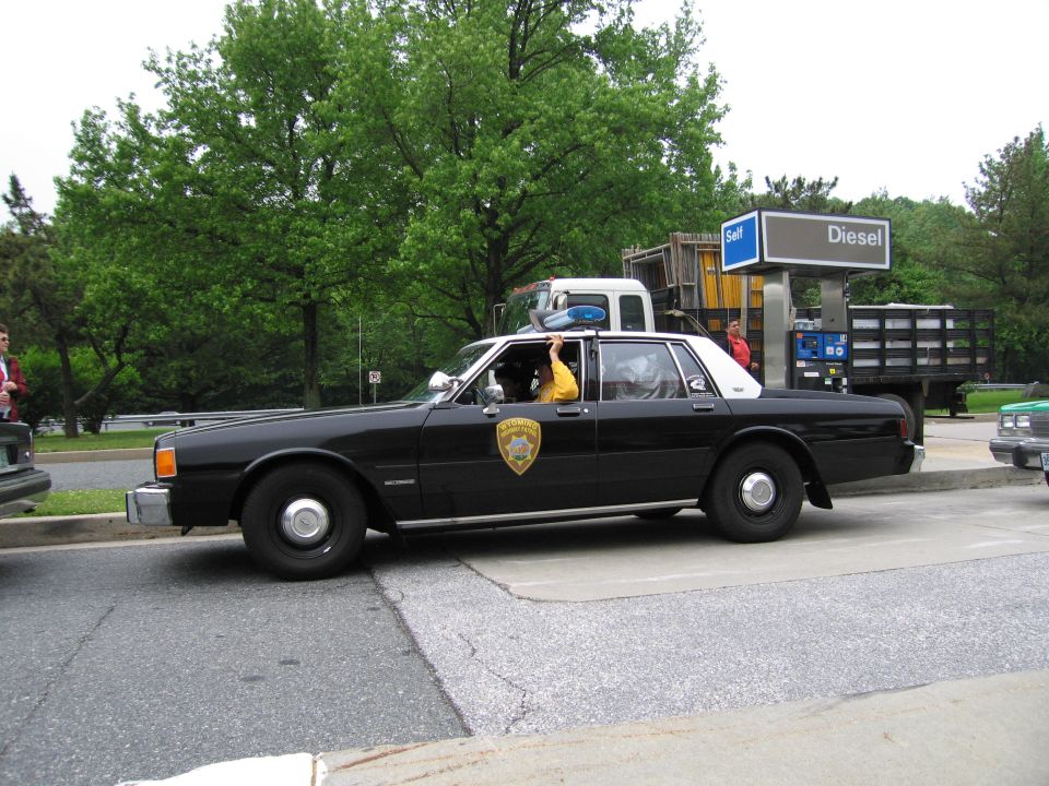 Black-and-white police sedan with occupants waving, driving past a Diesel fuel station