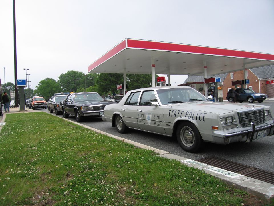 Tan State Police sedan parked at gas station with row of vintage police cars
