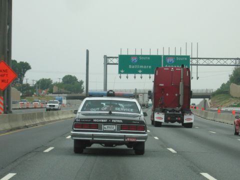 1991 Ford Crown Victoria - Police car with 'OUT OF SERVICE' sign on rear window, on highway behind a semi-truck.
