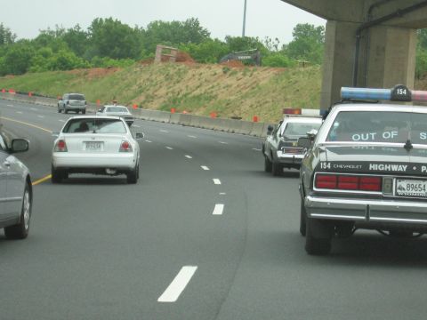 1991 Ford Crown Victoria - A police car with a light bar is seen from behind on a highway, with other vehicles in the distance.