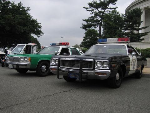 1991 Ford Crown Victoria - Two vintage police cars, a black and white and a green one, are parked side-by-side on pavement.