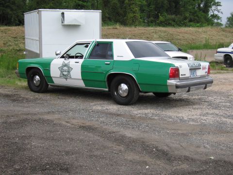 1991 Ford Crown Victoria - A green and white sheriff's car is parked next to a trailer, with another vehicle visible in the background.