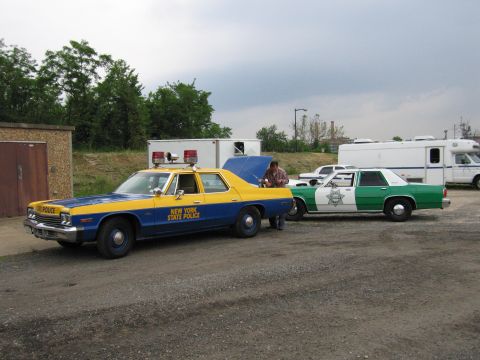 1991 Ford Crown Victoria - Two police cars, one blue and yellow and another green and white, are parked side-by-side on a gravel lot.