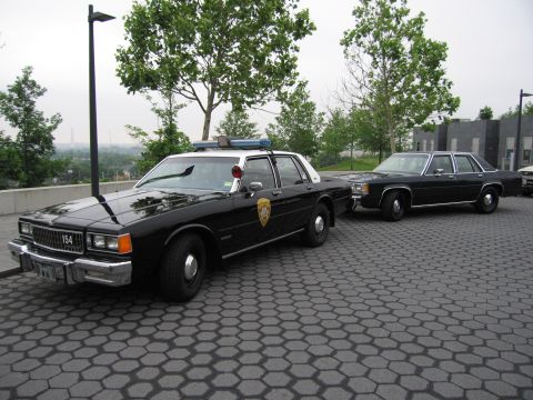 1991 Ford Crown Victoria - Two sedans, one a police car, are parked on a patterned pavement area with trees and buildings in the background.