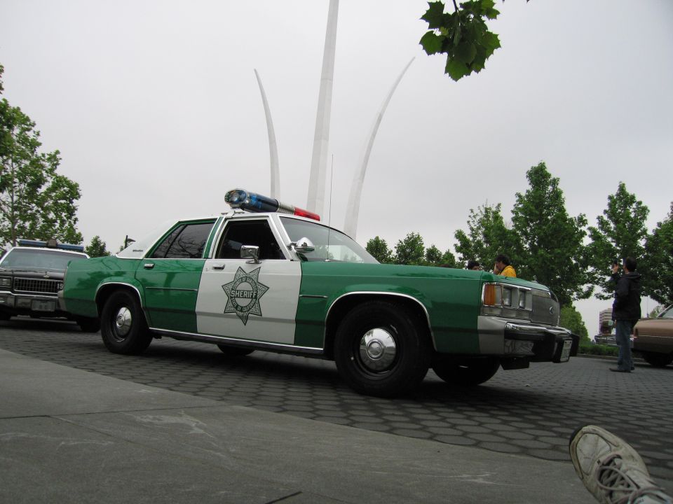 Green and white Ford Sheriff sedan, front three-quarter view, before arcing memorial spires
