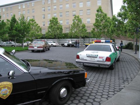 1991 Ford Crown Victoria - Multiple police vehicles are parked in a lot outside a large building.