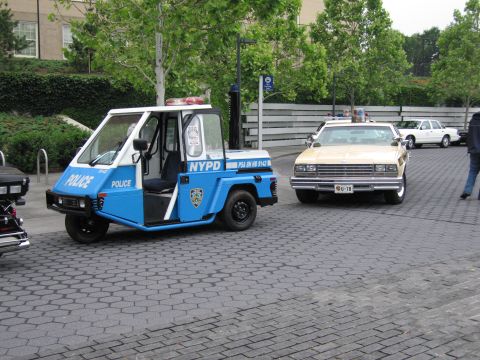 1991 Ford Crown Victoria - A blue and white NYPD three-wheeled vehicle is parked next to a tan police car on a paved area.