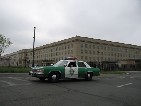 1991 Ford Crown Victoria - Police car with light bar parked in front of the Pentagon building under a cloudy sky.