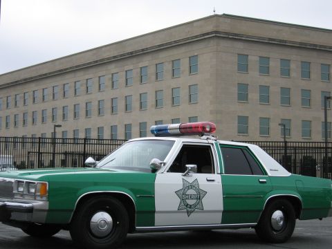 1991 Ford Crown Victoria - A sheriff's car with a star badge on the door is parked in front of a large, institutional-looking building.
