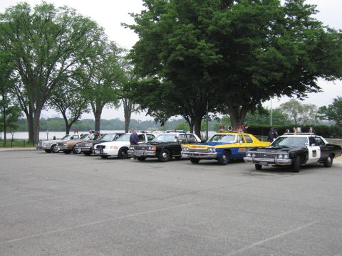 1991 Ford Crown Victoria - Several police vehicles are parked in a lot near a body of water and trees.