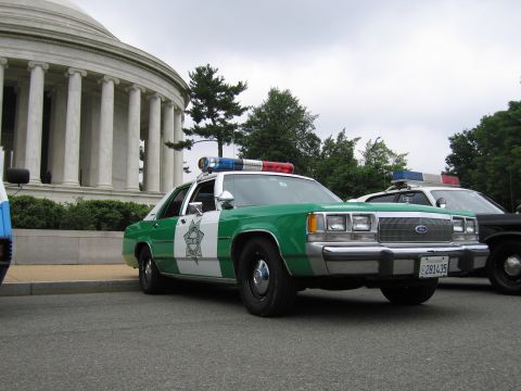 1991 Ford Crown Victoria - A police car with a light bar is parked in front of a large building with columns.