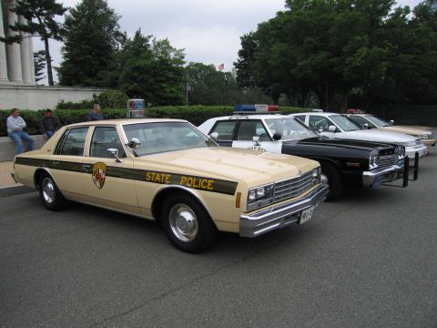 1991 Ford Crown Victoria - A line of police cars, including a tan 1977 model with "STATE POLICE" markings, are parked outdoors.