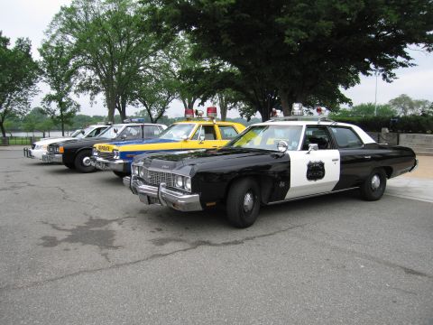 1991 Ford Crown Victoria - A line of police cars, including a black and white model with a shield emblem on the door, are parked outdoors.