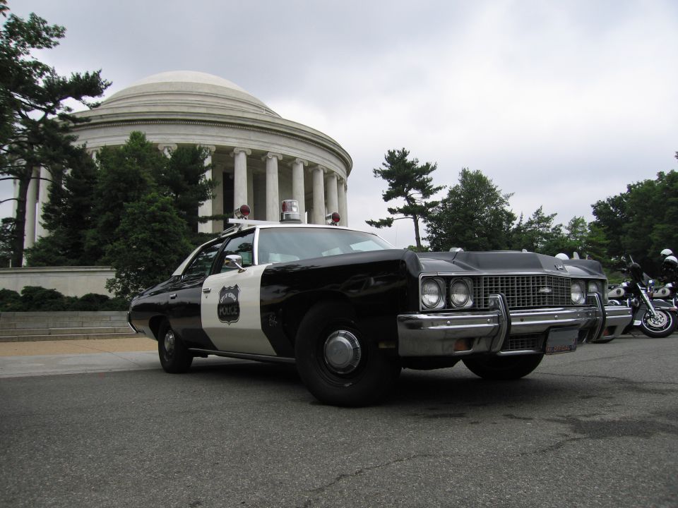 Black-and-white Chevrolet police cruiser parked before a domed memorial with motorcycle nearby