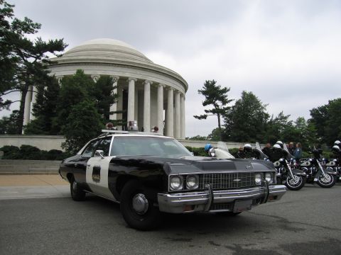 1991 Ford Crown Victoria - A vintage police car is parked in front of a large domed building, with a line of motorcycles and officers behind it.