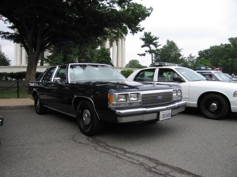1991 Ford Crown Victoria - Two police cars, one black and one white, are parked outdoors near a building with columns.