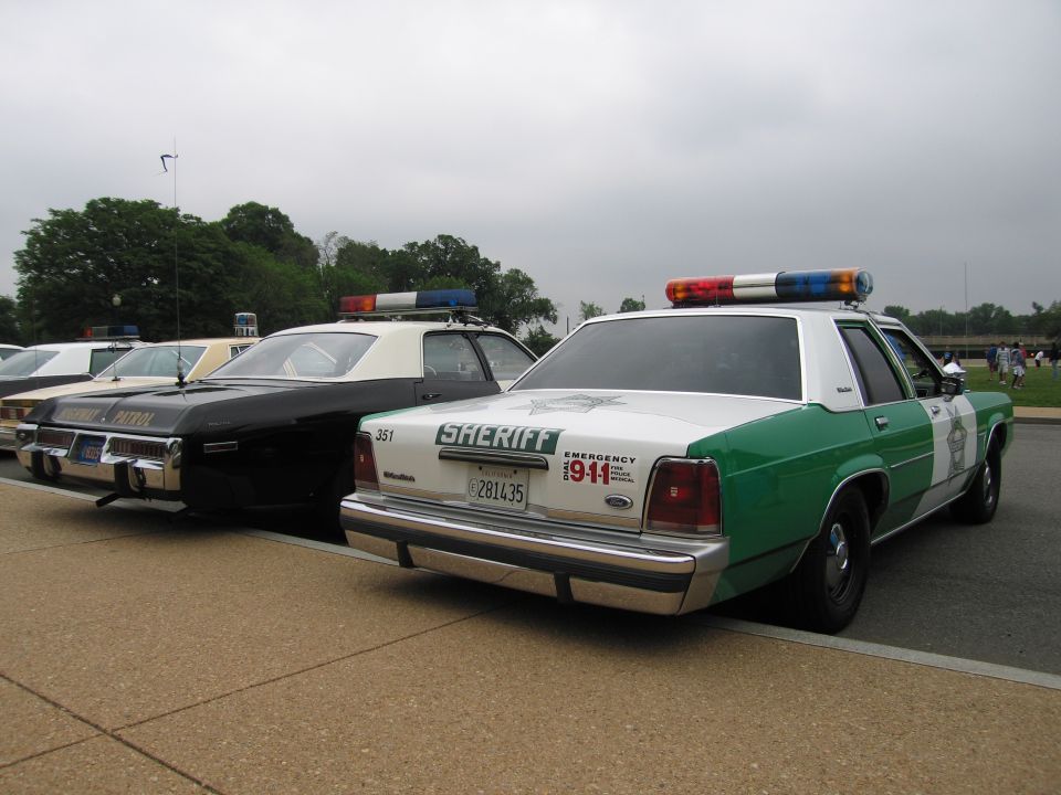 Green-and-white Ford Sheriff sedan with 911 markings beside black-and-white cruiser