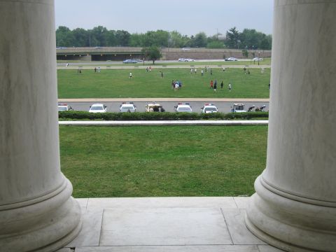 1991 Ford Crown Victoria - A line of police vehicles is parked on a road bordering a large green lawn with people scattered across it.