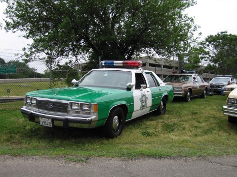 1991 Ford Crown Victoria - A vintage police car, likely from the 1980s, is parked on grass next to other vehicles.