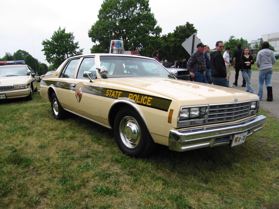 Tan State Police Chevrolet sedan parked on grass with onlookers standing nearby