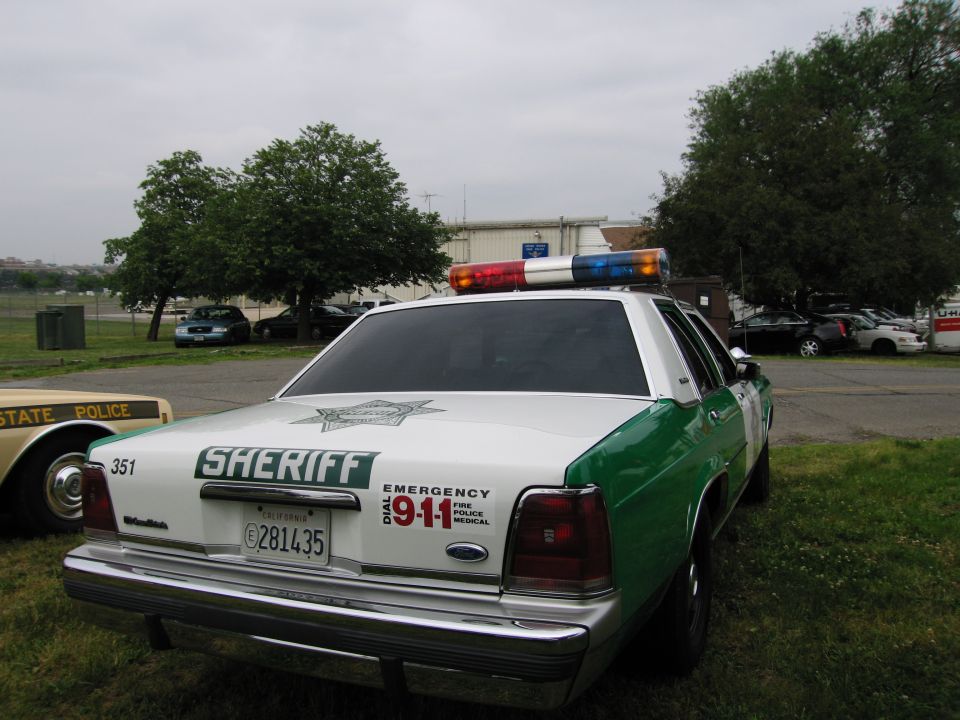 Rear of green-and-white Ford Sheriff sedan with 911 and 351 markings on grassy lot