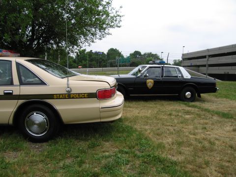 1991 Ford Crown Victoria - A tan and a black police car are parked side-by-side on a grassy area near a parking garage.