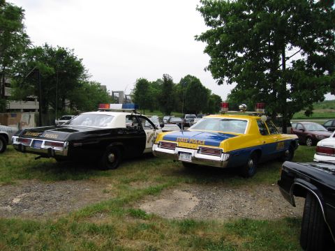 1991 Ford Crown Victoria - Two older model police cars are parked side-by-side on a gravel lot under a cloudy sky.