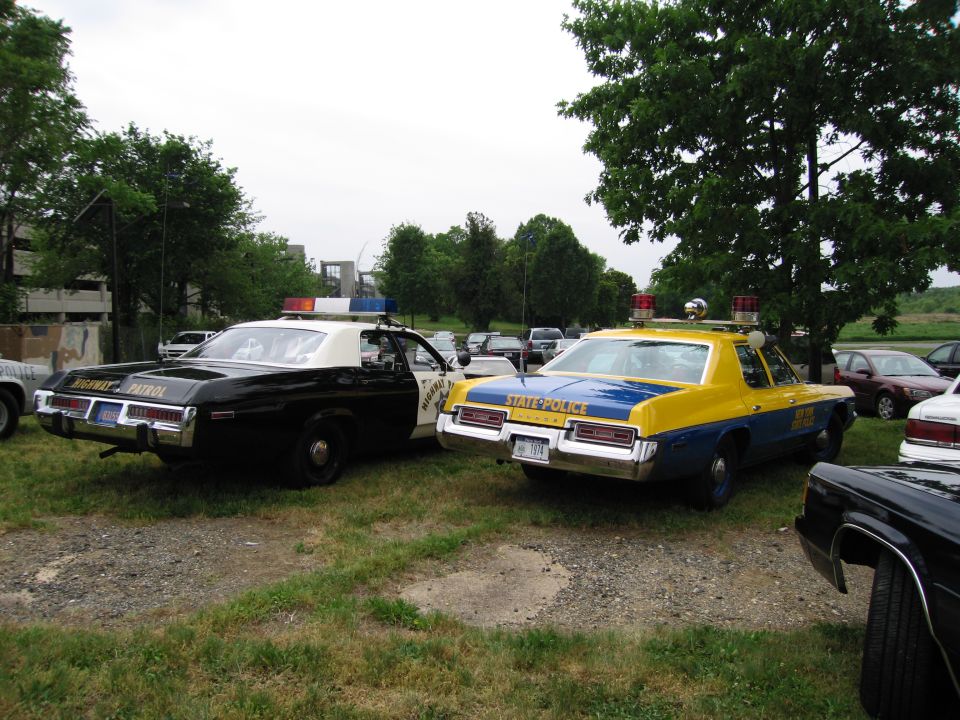 Black-and-white Highway Patrol car beside yellow-and-blue State Police sedan on grass