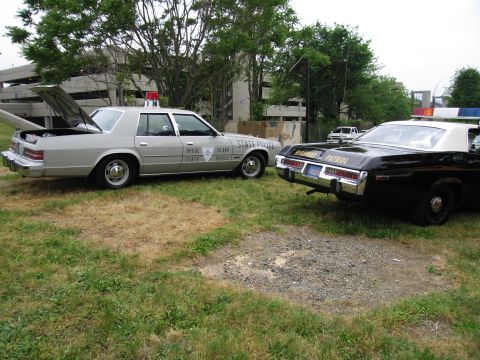 1991 Ford Crown Victoria - Two vintage police cars, a silver Rhode Island State Police vehicle and a black Highway Patrol car, are parked on a grassy area.