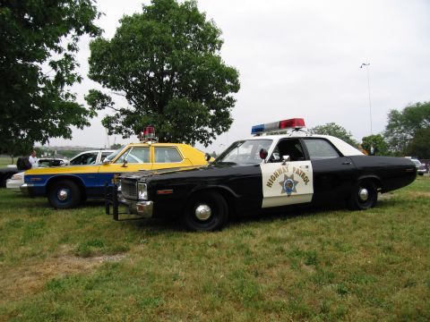 1991 Ford Crown Victoria - Two vintage police cars are parked side-by-side on a grassy field under trees.