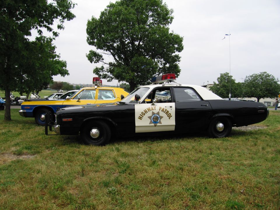 Black-and-white Highway Patrol sedan with twin roof lights parked on grass beside a yellow police car