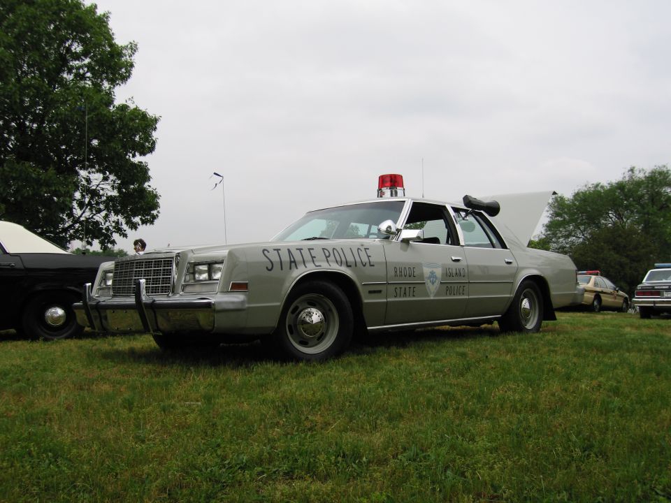 Silver Rhode Island State Police sedan with red roof beacon parked on grass, front three-quarter view