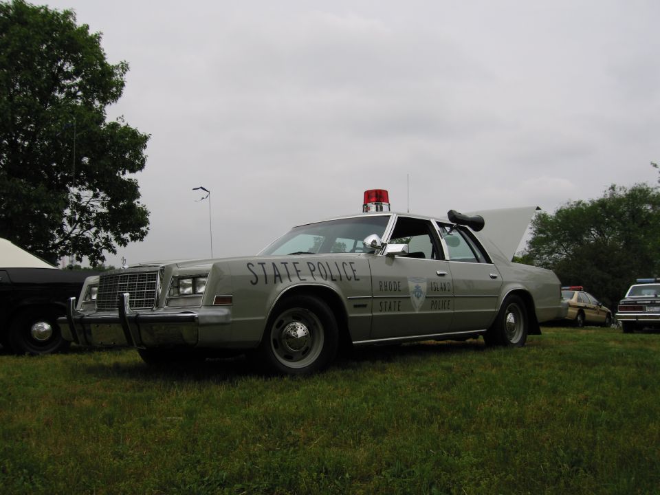 Silver Rhode Island State Police sedan with red beacon and spotlight parked on grass