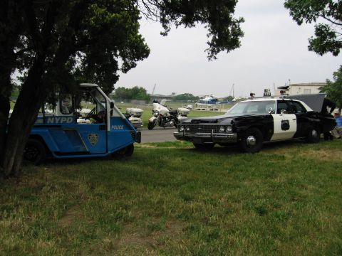 1991 Ford Crown Victoria - A black and white police car and a blue NYPD utility vehicle are parked on grass with other vehicles and helicopters in the distance.