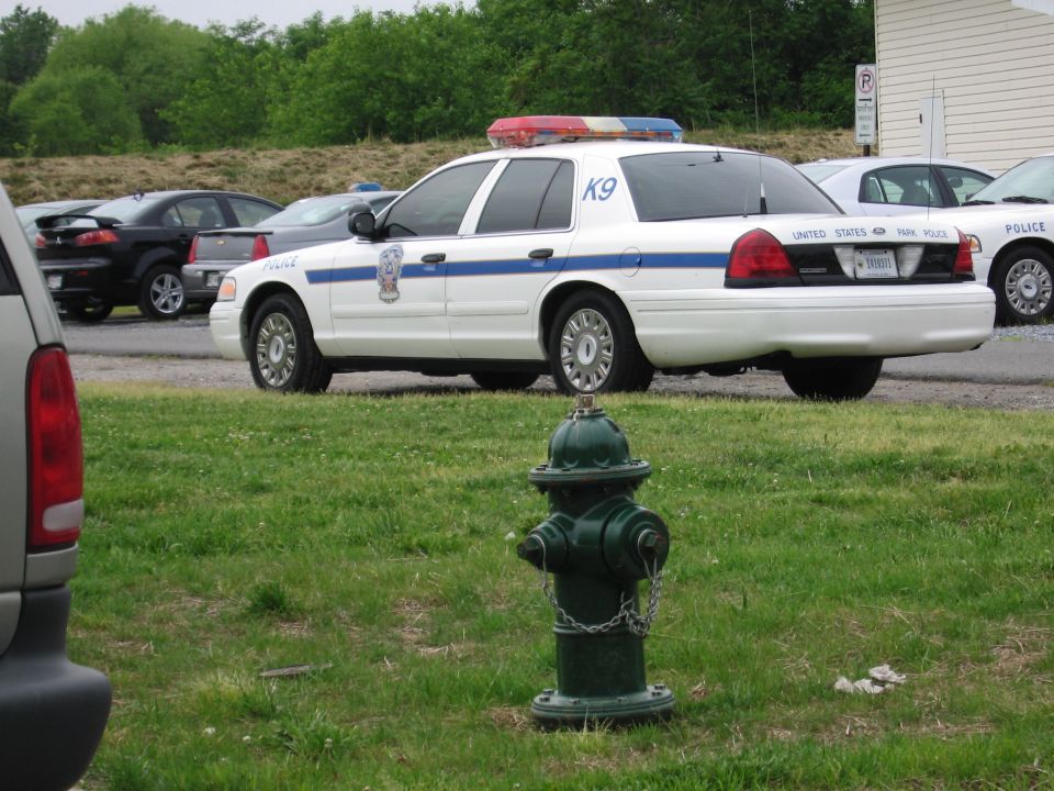 White US Park Police K9 cruiser parked behind a green fire hydrant beside an SUV on grass