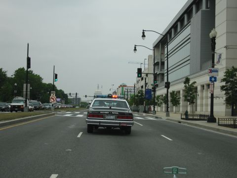 1991 Ford Crown Victoria - Police car on a city street, with traffic and buildings in the background. The Capitol dome is visible in the distance.