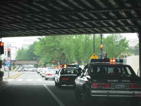 1991 Ford Crown Victoria - A line of police cars, including a 1991 Ford Crown Victoria, drives down a street under an overpass.