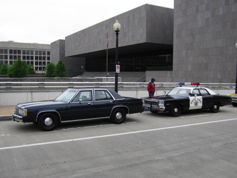 1991 Ford Crown Victoria - A dark sedan and a police car are parked on a street in front of a large, modern building.