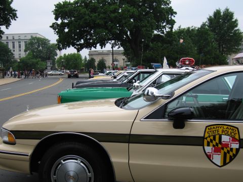 1991 Ford Crown Victoria - A line of police vehicles, including a tan car with a Maryland State Police emblem, are parked on a street with buildings and trees in the…