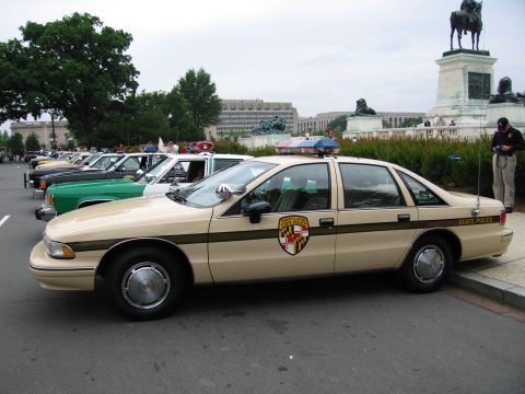 1991 Ford Crown Victoria - A line of police cars, including a tan 1991 Ford Crown Victoria, are parked in front of a monument.