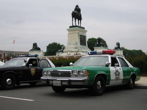 1991 Ford Crown Victoria - Two police cars are parked in front of a monument with lion statues and a statue of a man on a horse.