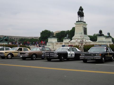 1991 Ford Crown Victoria - A line of vintage police cars, including a tan car with "STATE POLICE" visible on its door, are parked in front of a monument.