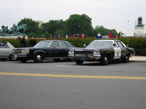 1991 Ford Crown Victoria - Two vintage police cars are parked on a street, with a monument and onlookers in the background.