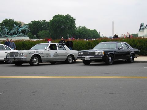 1991 Ford Crown Victoria - A Rhode Island State Police car and a black sedan are parked side-by-side with a monument in the background.