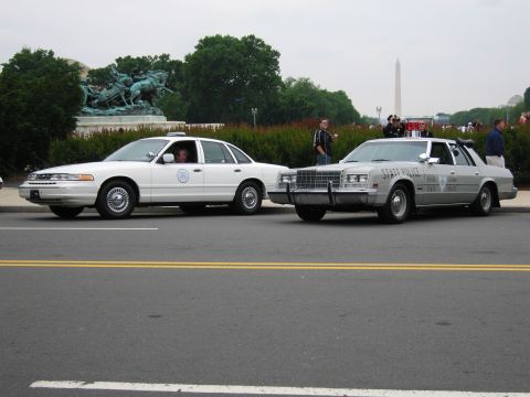 1991 Ford Crown Victoria - Two police cars, a white 1991 Ford Crown Victoria and a silver State Police vehicle, are parked on a street with monuments in the backgroun…