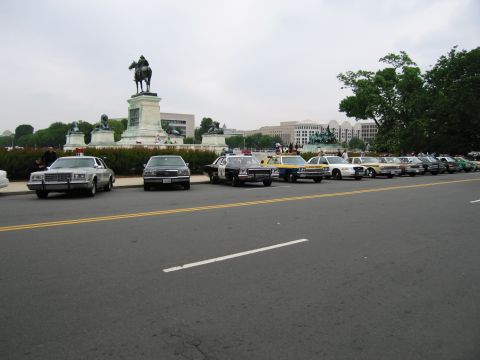 1991 Ford Crown Victoria - A row of vehicles, including police cars, are parked along a street with a monument and buildings in the distance.