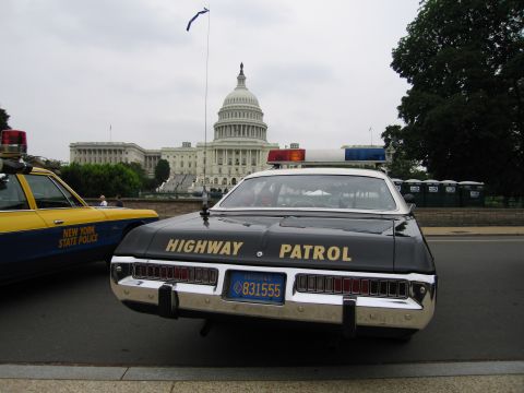 1991 Ford Crown Victoria - A black highway patrol car is parked next to a New York State Police vehicle with the Capitol building in the background.