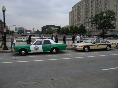 1991 Ford Crown Victoria - Two police cars are parked side-by-side on a street with pedestrians and buildings in the background.