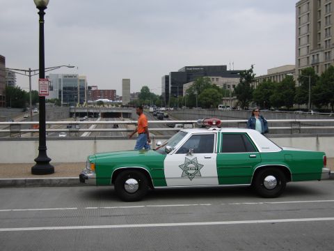 1991 Ford Crown Victoria - A two-tone green and white police car is parked on a bridge with city buildings in the background.