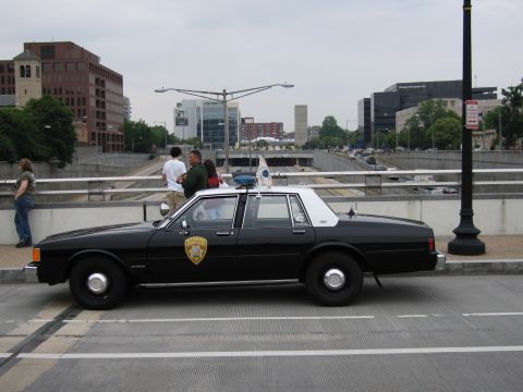 1991 Ford Crown Victoria - A 1991 Ford Crown Victoria police car is parked on the side of a road with a city skyline in the background.